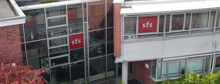 Drohnenaufnahme der Rückseite der Sozialforschungsstelle Dortmund an einem sonnigen Herbsttag. Hierbei ist an zwei Fenstern das rote Logo der sfs, ausgeschrieben in weißen Buchstaben, zu sehen. 