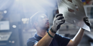A man with safety goggles and gloves holds a sheet of metal