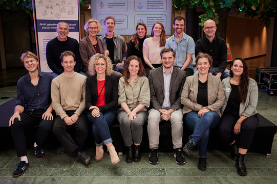 Gruppenfoto: 7 Personen sitzen auf einem Sofa, dahinter stehen 7 weitere Personen. 