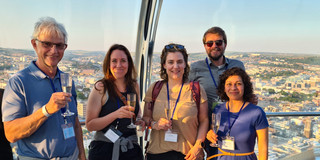 Foto: Gruppe von Personen steht mit Getränken vor einer Aussicht über eine Stadt.