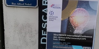 Ein Schild vor einem Gebäude zeigt die Aufschrift „Institut Français d’Urbanisme“ und „Cité Descartes“. Darunter ist ein Plakat mit Informationen zu einer internationalen Konferenz angebracht. Links daneben befindet sich ein Straßenschild mit der Adresse „2 Rue Alfred Nobel“.