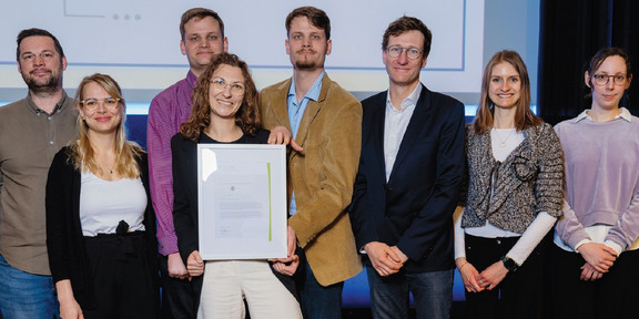 Gruppenfoto von acht Personen. Zwei Personen halten eine gerahmte Urkunde.