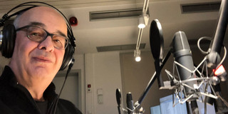 Foto: Person mit Kopfhörern an einem Tischmikrofon in einem Aufnahmeraum; mehrere Studiomikrofone, Popschutz und Monitore sichtbar.