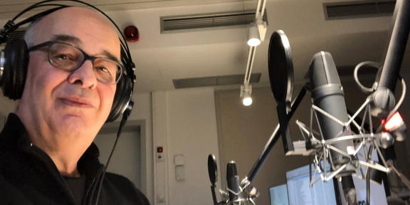 Foto: Person mit Kopfhörern an einem Tischmikrofon in einem Aufnahmeraum; mehrere Studiomikrofone, Popschutz und Monitore sichtbar.