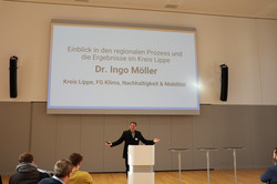 Dr. Ingo Möller steht an einem Rednerpult und macht eine Geste. Hinter ihm wird eine Präsentation auf eine Leinwand projiziert. Einige Zuhörer sitzen ein paar Meter von ihm entfernt.