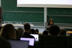 Foto: Eine Person sitzt in einem Hörsaal vor einer Tafel mit mathematischen Formeln. Mehrere Personen im Publikum folgen der Veranstaltung, einige arbeiten an Laptops.