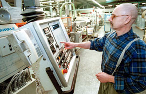 Ein Mann mit blau – schwarz kariertem Hemd und Brille steht vor einem Bedienfeld einer Maschine mit leuchtenden Knöpfen im unten Bereich. Er liest den Text rechts über den Knöpfen. Links an dem Bedienfeld ist ein Telefon befestigt.
