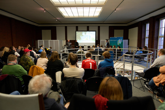 Foto: Mehrere Personen sitzen in einem Raum und hören einer Präsentation zu. Vorne befindet sich eine Projektionsfläche mit einer Leinwand und mehreren Vortragenden.