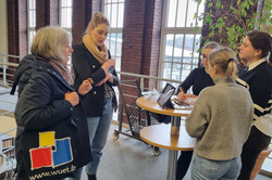 Foto: Personen stehen an einem Stehtisch und unterhalten sich in einem Gebäude mit großen Fenstern.