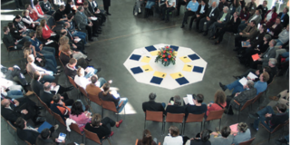 Viele Personen sitzen in einem großen Kreis in einem hellen Raum. In der Mitte liegen farbige Tafeln und ein Blumenstrauß auf dem Boden