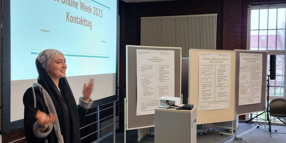 Foto: Person gestikuliert vor einer Projektion mit dem Titel "Get Online Week 2023 Kontakttag"; mehrere mobile Stellwände mit Informationsplakaten und ein Präsentationspult mit Projektor sind vorhanden.