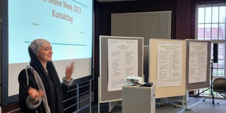Foto: Person gestikuliert vor einer Projektion mit dem Titel "Get Online Week 2023 Kontakttag"; mehrere mobile Stellwände mit Informationsplakaten und ein Präsentationspult mit Projektor sind vorhanden.