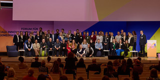 oto: Eine große Gruppe von Personen steht und sitzt auf einer Bühne während einer Veranstaltung. Auf der Leinwand im Hintergrund steht „Vielen Dank!“. Im Vordergrund sitzen Zuschauerinnen und Zuschauer.