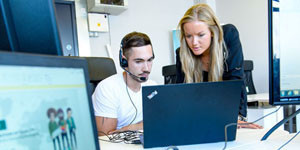 Werden sich durch den Wandel der Arbeit die Geschlechterverhältnisse verändern? Büroschreibtisch. Mann sitzt mit Headset an seinem Laptop. Frau steht mit linker Hand am Tisch angelehnt neben ihm, schaut auf den Laptop. Im Vordergrund verschwommen ein Laptop mit Kabelverbindung zu einem PC-Bildschirm.