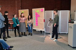 Foto: Kleine Gruppe in einer Workshop-Situation; mehrere Teilnehmende stehen an den Pinnwänden, ein Präsentierender mit Laptop spricht, weitere Teilnehmende sitzen an der Seite.