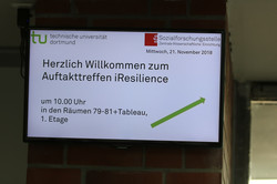 Foto eines Bildschirms, auf dem eine Präsentation gezeigt wird. Neben den Logos der TU Dortmund und der sfs steht dort das Datum "Mittwoch, 21. November 2018". Darunter steht "Herzlich Willkommen zum Auftakttreffen iResilience" und "um 10.00 Uhr in den Räumen 79-81+Tableu, 1. Etage".