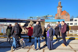 Foto: Eine Gruppe von Menschen steht auf einem Platz am Hafen in Dortmund und hört einer Person zu, die offenbar etwas erklärt.