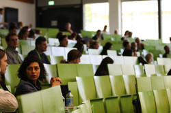 Personen sitzen in einem Hörsaal mit grünen Sitzreihen und hören einer Veranstaltung zu.