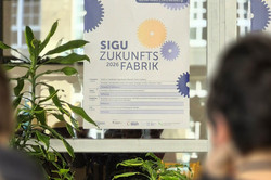 Photo: People sitting with their backs to the camera are looking at a poster for SIGU Zukunftsfabrik 2026.