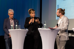 Podiumsdiskussion 3 Personen stehen auf einer Bühne mit 2 Stehtischen