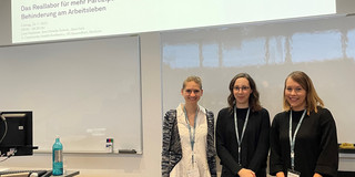 Foto: Drei Personen stehen in einem Seminarraum vor einer Leinwand, auf der eine Präsentationsfolie mit dem Titel „Das Reallabor für mehr Partizipation und Empowerment von Menschen mit Behinderung am Arbeitsleben“ und das Logo der Technischen Universität Dortmund zu sehen ist.