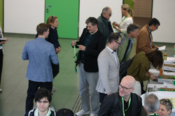 Foto: Teilnehmende einer Veranstaltung bewegen sich zwischen Tischen, füllen Formulare aus oder lesen Unterlagen; mehrere tragen Namensschilder und stehen in kleinen Gesprächsgruppen.
