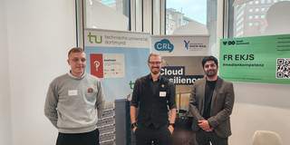 Drei Personen stehen in einem Raum vor mehreren Roll-up-Bannern, auf denen unter anderem das Logo der Technischen Universität Dortmund und der Schriftzug „Cloud Resilience“ zu sehen sind.