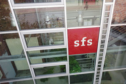 Drohnenaufnahme der verglasten Rückseite der Sozialforschungsstelle Dortmund an der Deutschen Straße in Eving mit einem roten sfs-Logo auf einem Fenster.