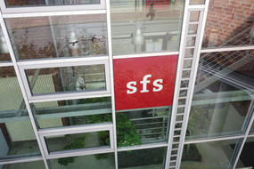 Drohnenaufnahme der verglasten Rückseite der Sozialforschungsstelle Dortmund an der Deutschen Straße in Eving mit einem roten sfs-Logo auf einem Fenster.