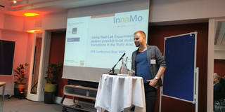 Kay Cepera stands at a standing desk with a standing microphone, behind him a presentation is shown on a screen