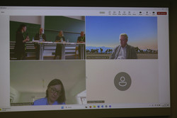 Foto: Eine Videokonferenz wird auf einem Bildschirm angezeigt, mit mehreren Teilnehmenden in verschiedenen Fenstern. In einem Fenster ist eine Podiumsdiskussion in einem Hörsaal zu sehen, in anderen Personen vor unterschiedlichen Hintergründen.