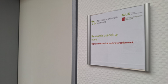 Photo: A door sign featuring the logos of TU Dortmund University, the Faculty of Social Sciences, and the sfs. Below the logos is the text “Research Assistant (m/f/d)” and “Work in the service work/interaction work”. Next to the door sign is a closed door.