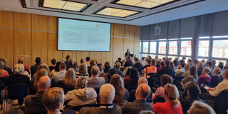 Lisa Preissner is standing in a hall where many people are seated in several rows of chairs. She is standing at a lectern with a laptop open in front of her. A presentation is being shown on a screen.