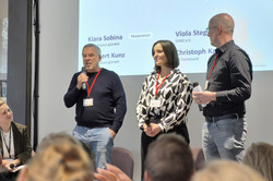 Photo: Four people in front of an audience; three are standing, and one is sitting. They are holding microphones and wearing ID badges around their necks.