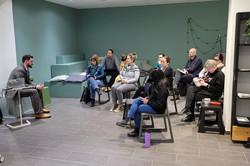 10 Personen sitzen im Halbkreis in zwei Reihen um eine Person. Manche machen sich Notizen.
