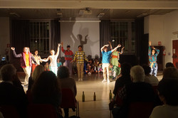 Foto einer Theateraufführung. Acht Personen stehen in einem mit Glasflaschen abgegrenzten Bereich und führen Bewegungen aus. Rundherum sitzen Personen und schauen ihnen zu.