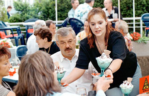 Menschen sitzen draußen an einem Eiscafé. Kellnerin bedient sie mit Eisbechern.