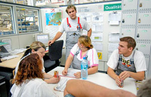 Meeting in a partitioned office with sheets of paper on the wall. Five employees in white shirts and gray dungarees sit at the table. Drinks are on the table. One employee is leaning on a chair behind them. Lockers in the background on the right, a desk with a computer screen in the background on the left.