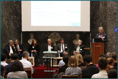 Social Innovation - Research and Policy of the Future towards a Comprehensive Innovation Policy Sechs Personen im Anzug sitzen auf einer Bühne. Ein Mann steht an einem Rednerpult und hält eine Präsentation vor Publikum.
